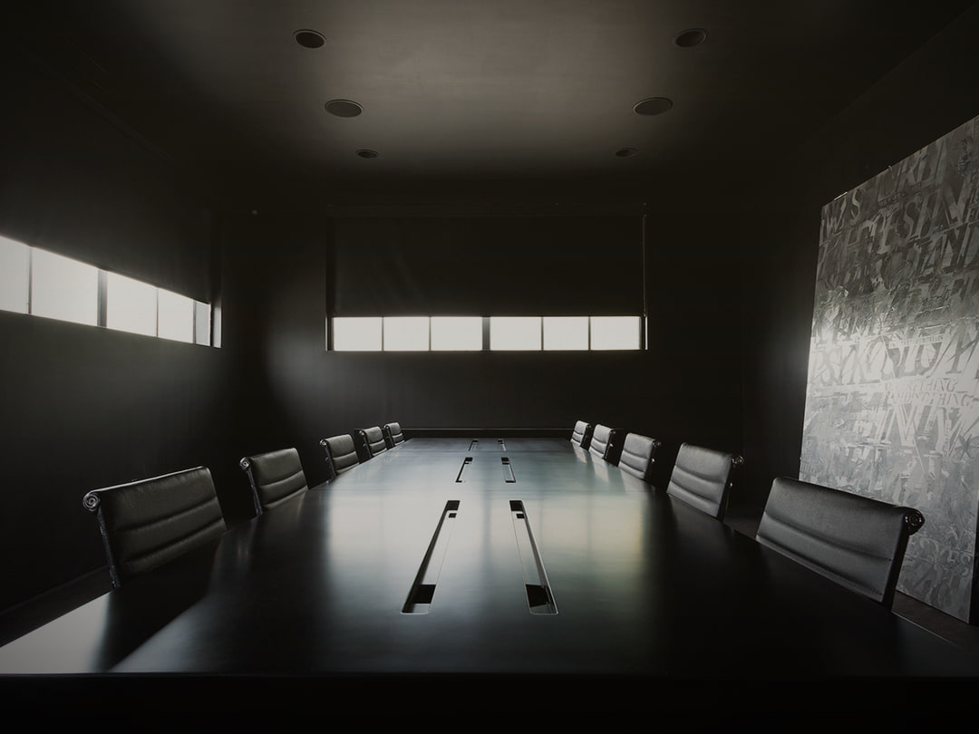 A stark private boardroom lit through horizontal blinds: scarcity governed behind closed doors